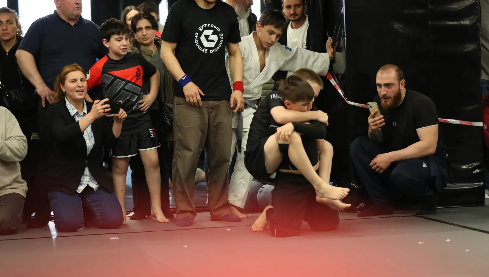 Parents and teammates crouched at the edge of the mat watching a match at a Gymnasia kids competition in Tbilisi